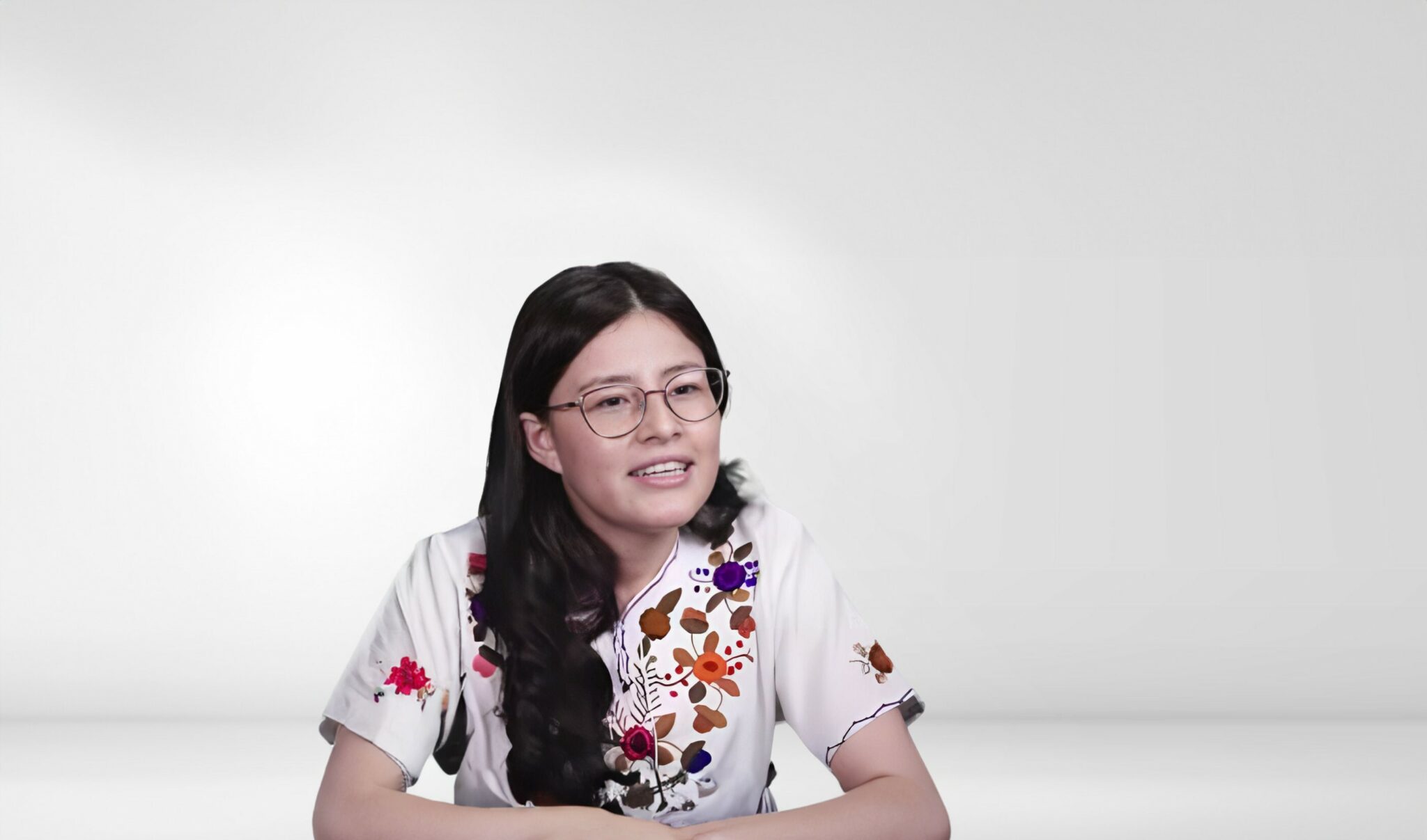 A woman with long hair and glasses sitting in front of a white wall.